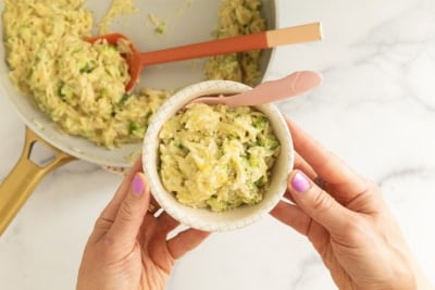 Hands holding a bowl of creamy broccoli orzo with a skillet in the background.