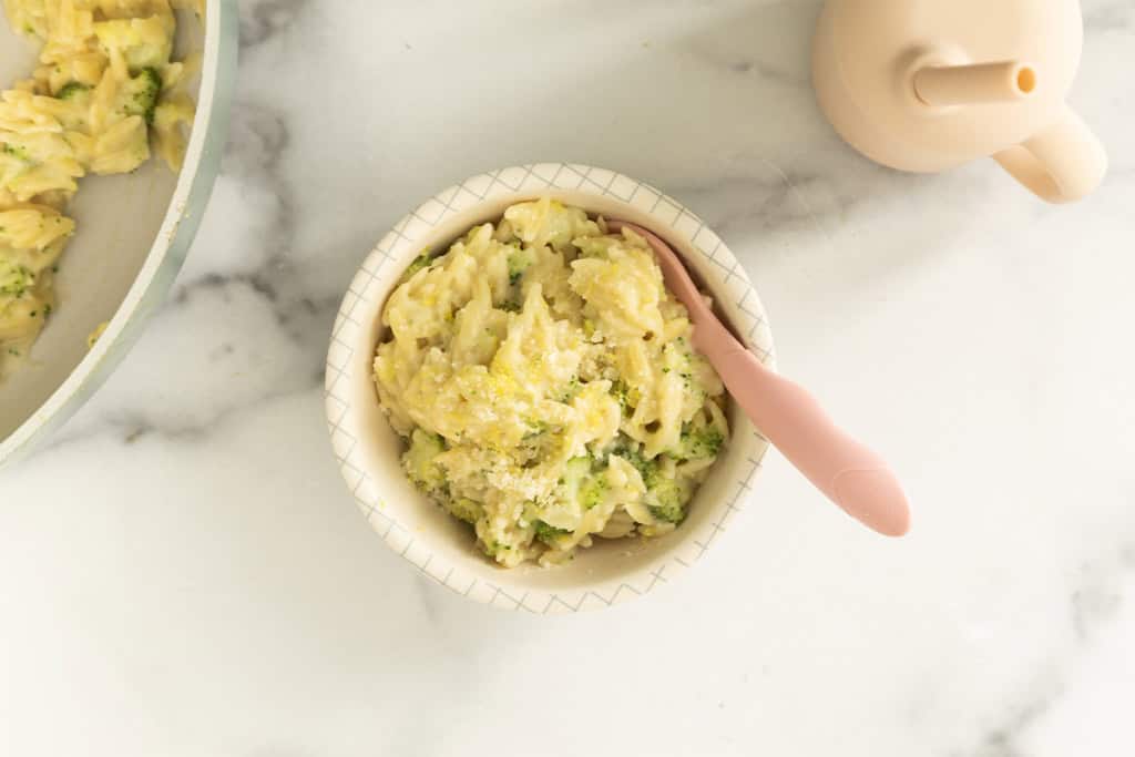 Bowl of creamy broccoli orzo with a pink spoon and skillet nearby.