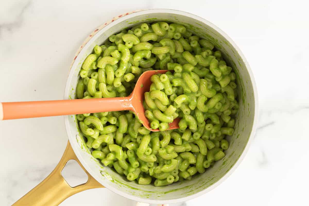 Green mac and cheese in a pot with an orange serving spoon.