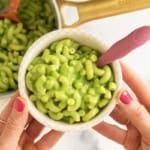 A bowl of green mac and cheese held over a pot of pasta.