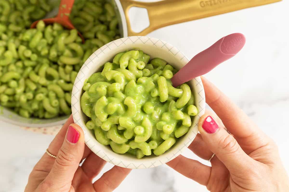 A bowl of green mac and cheese held over a pot of pasta.
