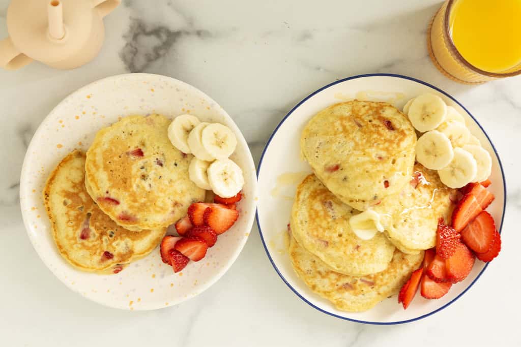 Strawberry banana pancakes on two plates with sliced bananas and strawberries.