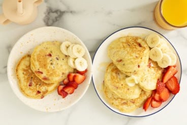 Strawberry banana pancakes on two plates with sliced bananas and strawberries.